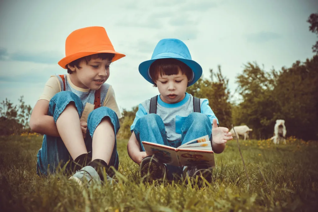 Two young boys enjoying a book together outdoors on a sunny day, wearing vibrant hats.